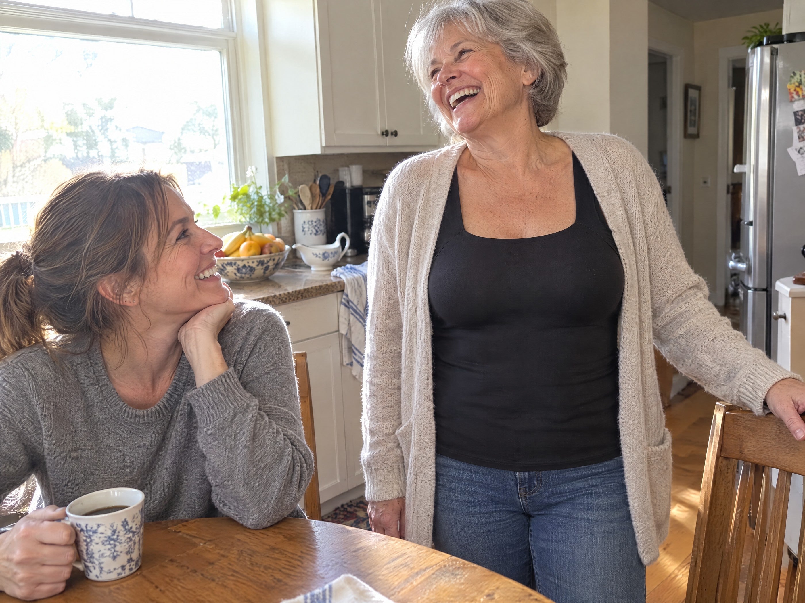 A woman of 64 laughing in her kitchen, a cardigan over a black tank, her daughter at the table